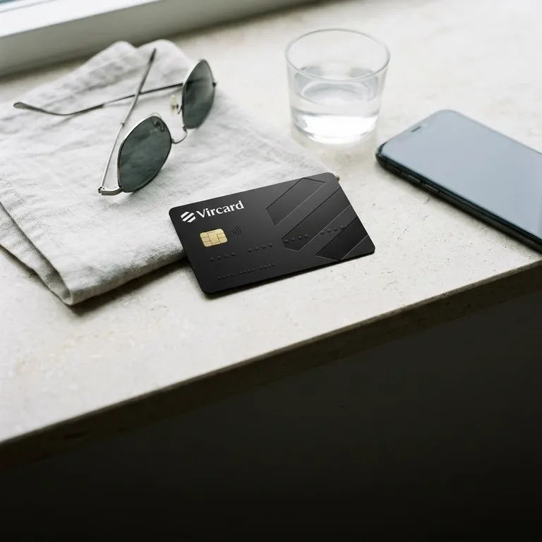 Чёрная карта Vircard на светлом подоконнике рядом с очками, стаканом воды и телефоном